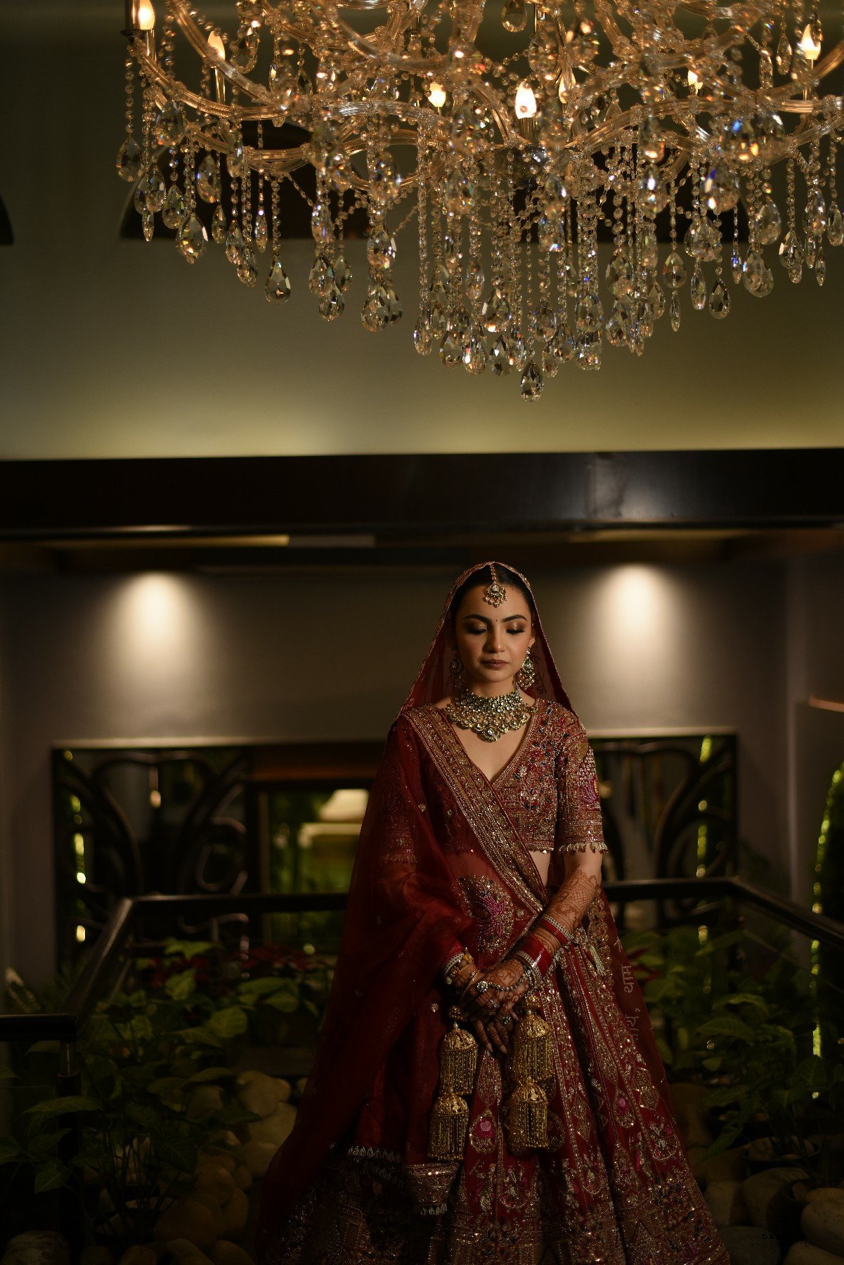 Indian bride in red lehenga under grand crystal chandelier at luxury reception, professional wedding photography